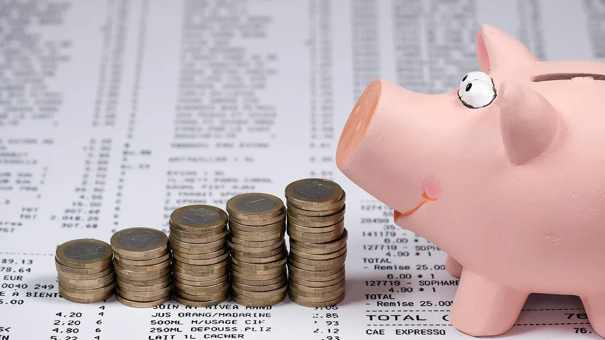 A pink piggy bank with a coin slot and a single coin inserted into it is positioned to the right, accompanied by four stacks of coins to its left, all situated on top of a receipt or invoice featuring black text on a white background.