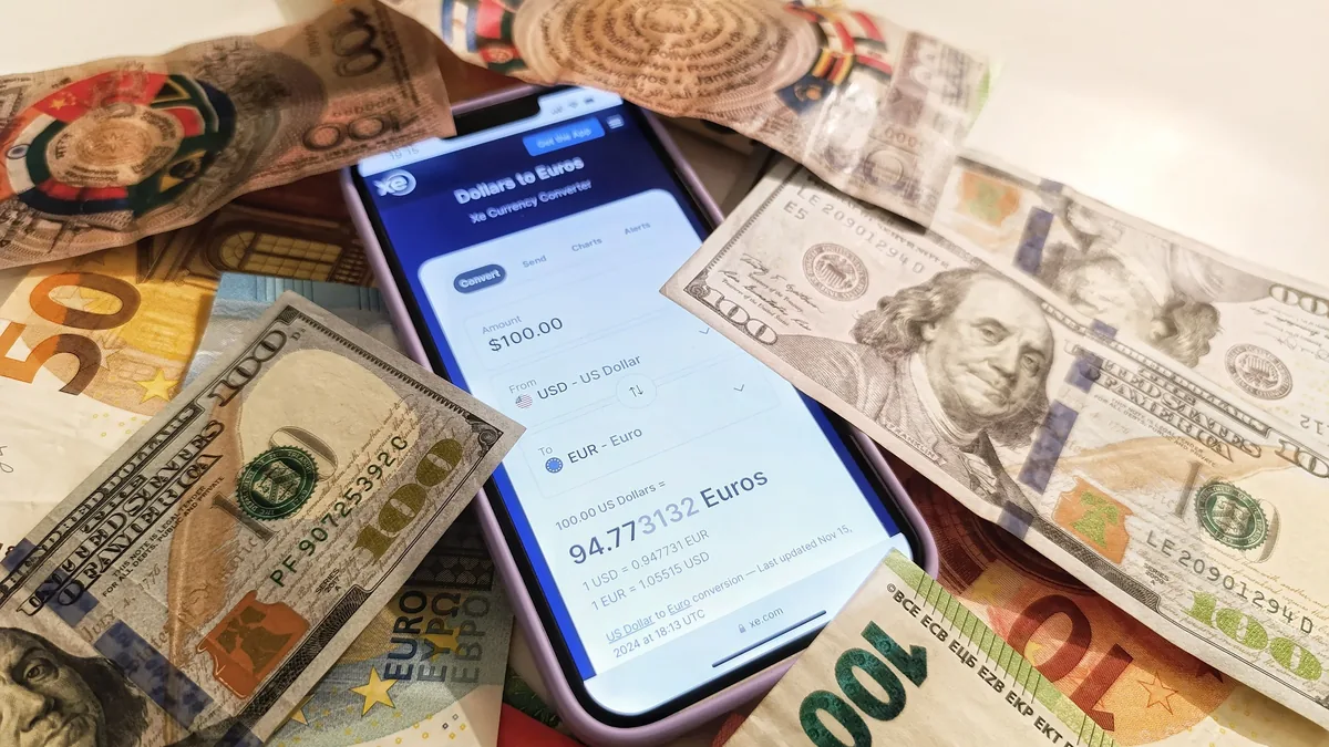 A smartphone displaying a currency exchange rate on a screen, surrounded by various denominations of US dollar and Euro banknotes.